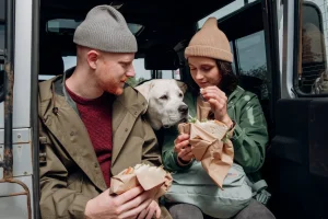 A man and woman sit in the back of a van, smiling with a dog resting between them.